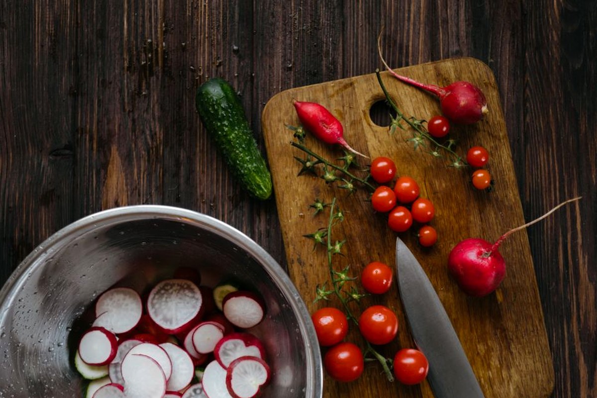 Fresh radishes, tomatoes, cucumber, and herbs prepared on a wooden kitchen table.